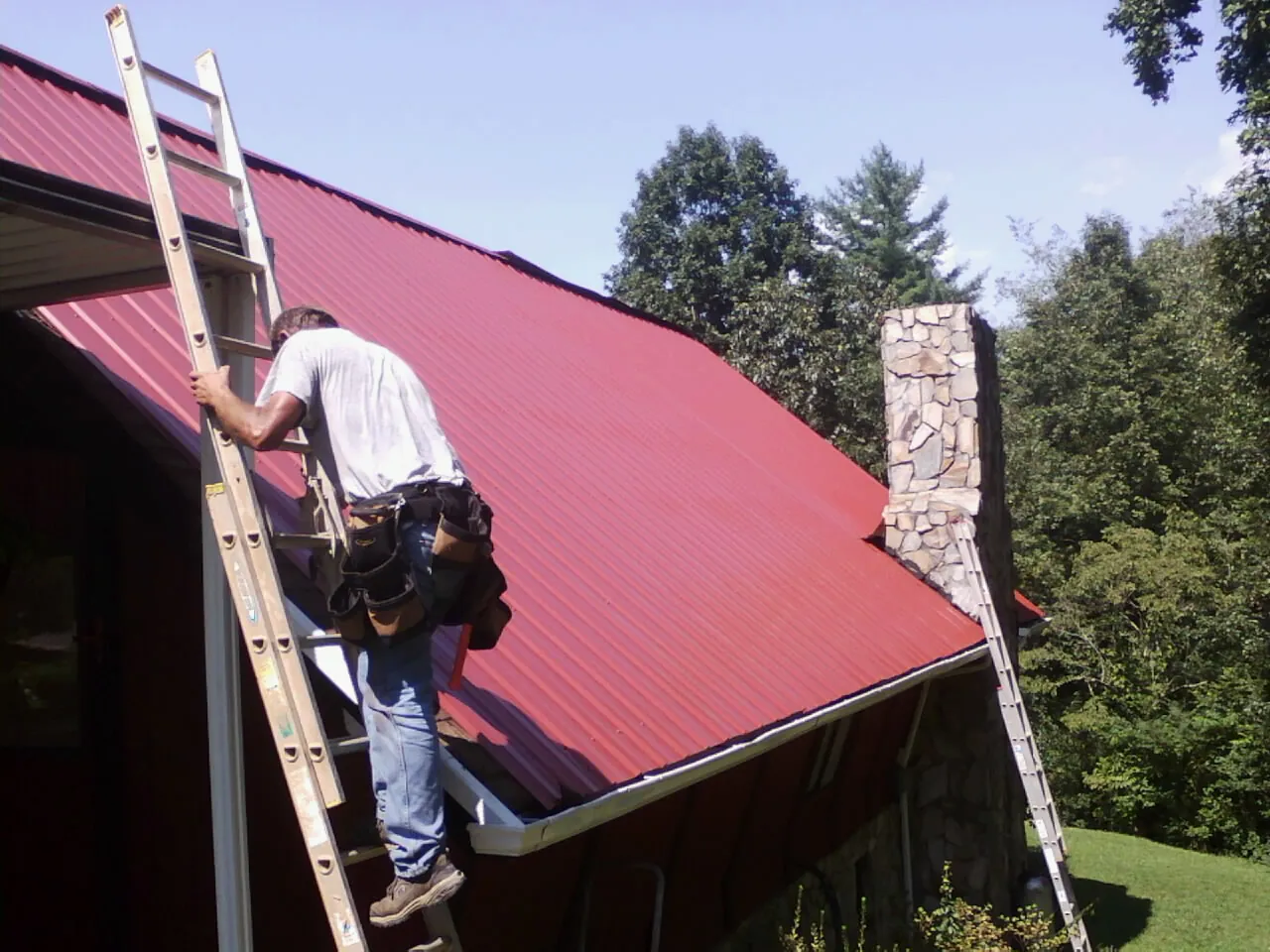 Roofer on a ladder beside a red metal roof and stone chimney — Soffit & Fascia Repair by Roofing Brothers in New Columbia